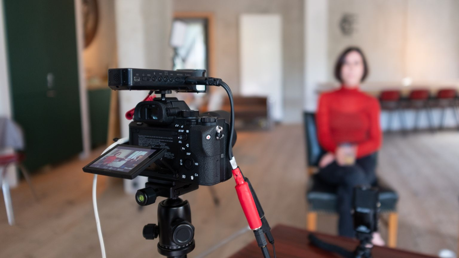 A video camera on a tripod records a person sitting in a chair for a Story Corner interview in a bright, modern room with wooden floors and natural light. The camera’s screen shows the person framed in the shot, suggesting a storytelling or filming setup.